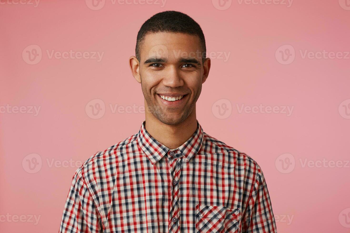 Close up of young happy attractive dark skinned guy in checkered shirt, looks at the camera with positive expression, broadly smiling and stands over pink background. photo