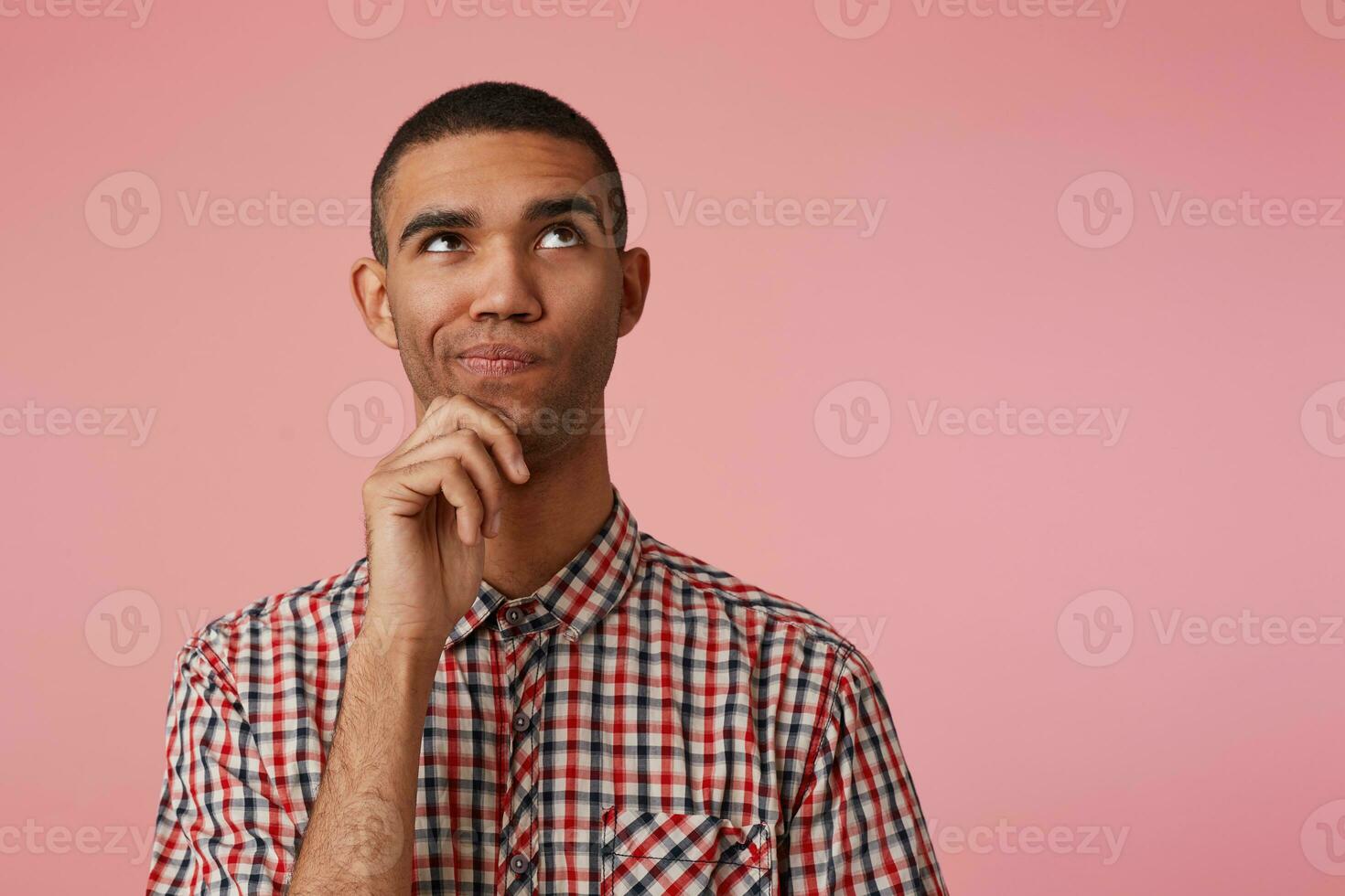 Close up of young pensive attractive dark skinned guy in checkered shirt, looks away and touches chin, thinks and doubts, stands over pink background with copy space at the right side. photo