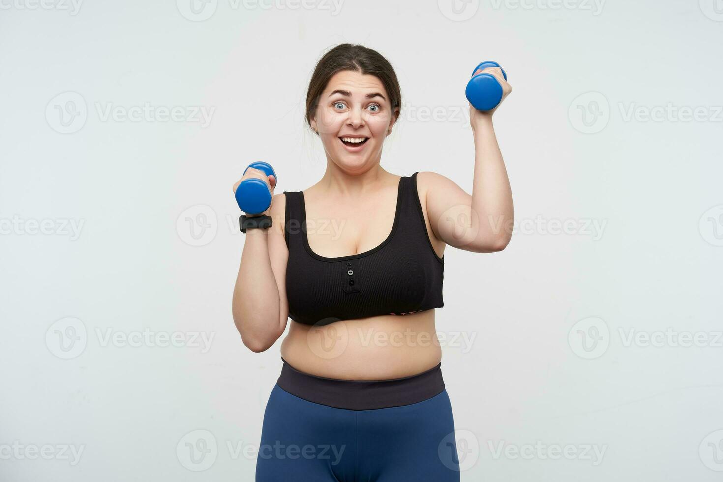 Overjoyed young brunette plump female dressed in sporty wear training her arms using little blue dumbbells while looking excitedly at camera, posing over white background photo