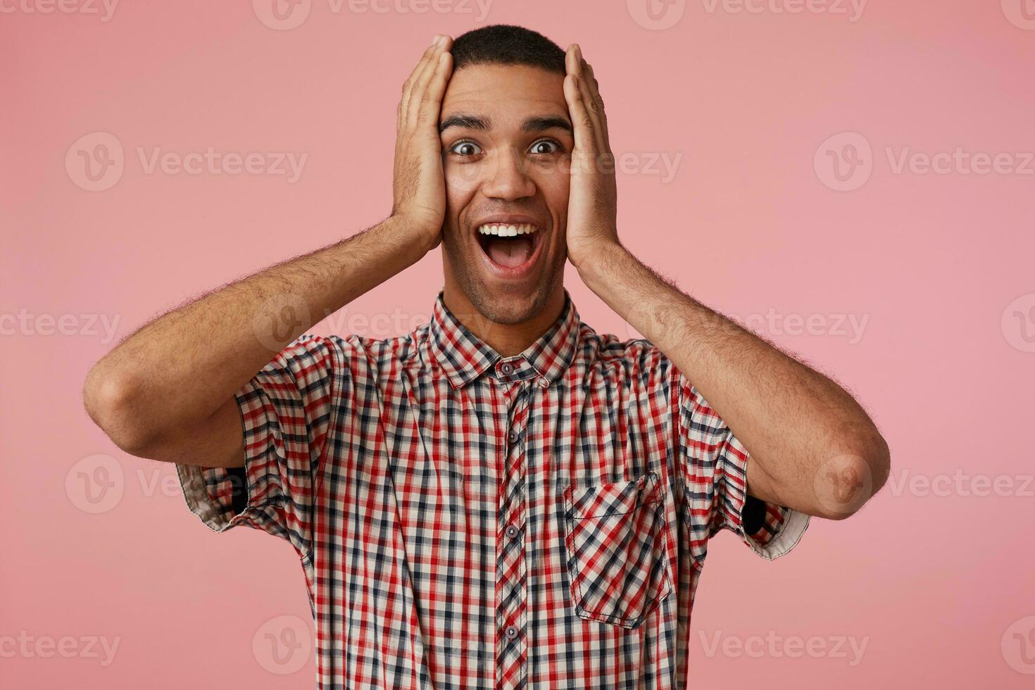 Wow Socked young attractive dark skinned guy won a car, looks at the camera with wide open mouth and eyes, stands over pink background and holding his head, wears checkered shirt. photo