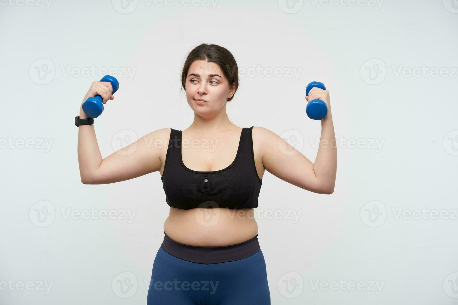 Pensive young oversized female with ponytail hairstyle making work out for hands and looking thoughtfully aside, posing over white background. Body positive concept photo