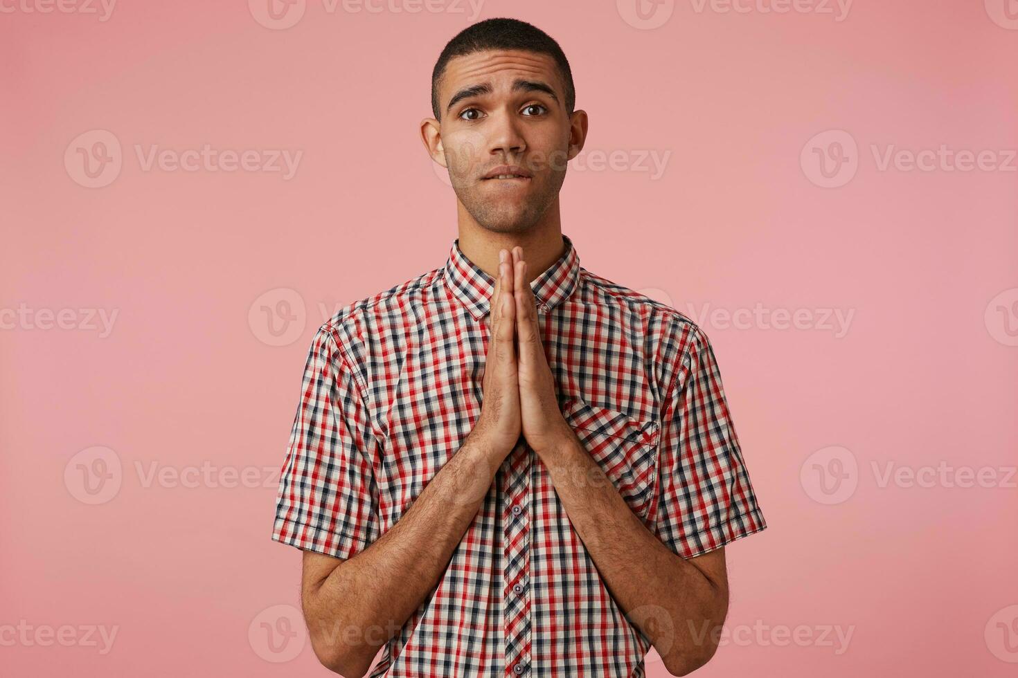 Portrait of young attractive dark skinned guy in checkered shirt, cupped hands together in pray gesture, bites lip, look at the camera and hopes for luck,stands over pink background. photo
