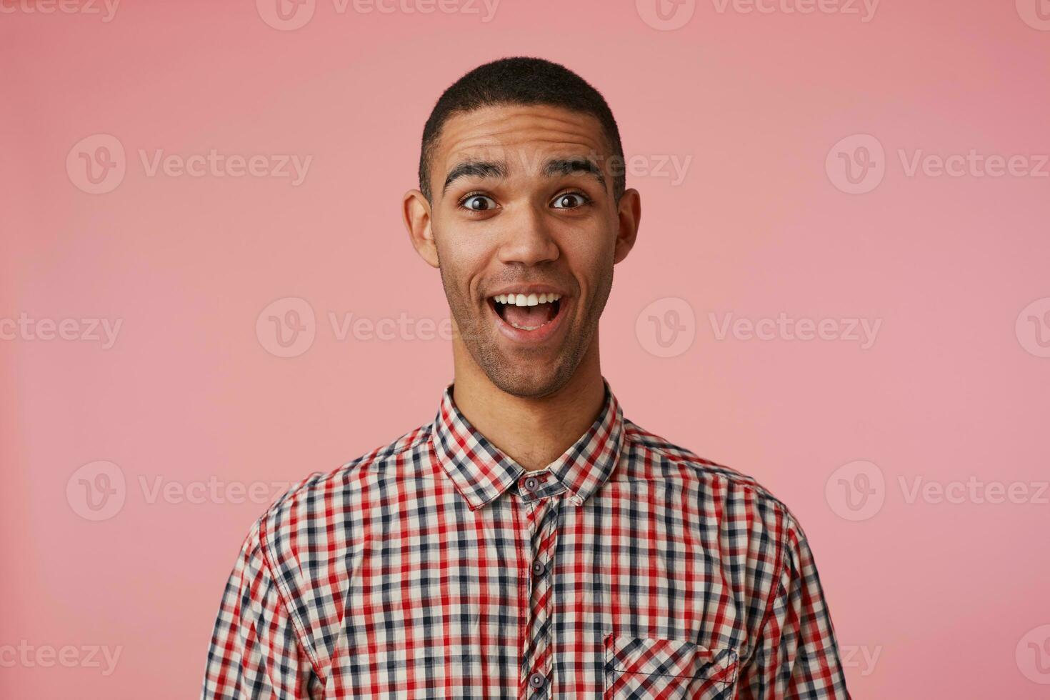 Portrait of happy amazed young attractive dark skinned guy in checkered shirt, looks at the camera with wide open mouth and eyes, stands over pink background with shoked expression. photo
