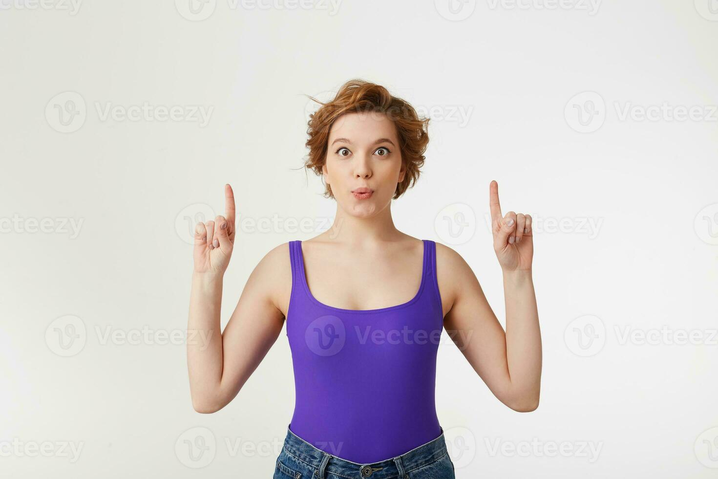 Portrait of a young happy amazed attractive short-haired girl, wearing a purple jersey, looking at the camera, speaks lips wow and index fingers point up at the copy space isolated over white wall. photo