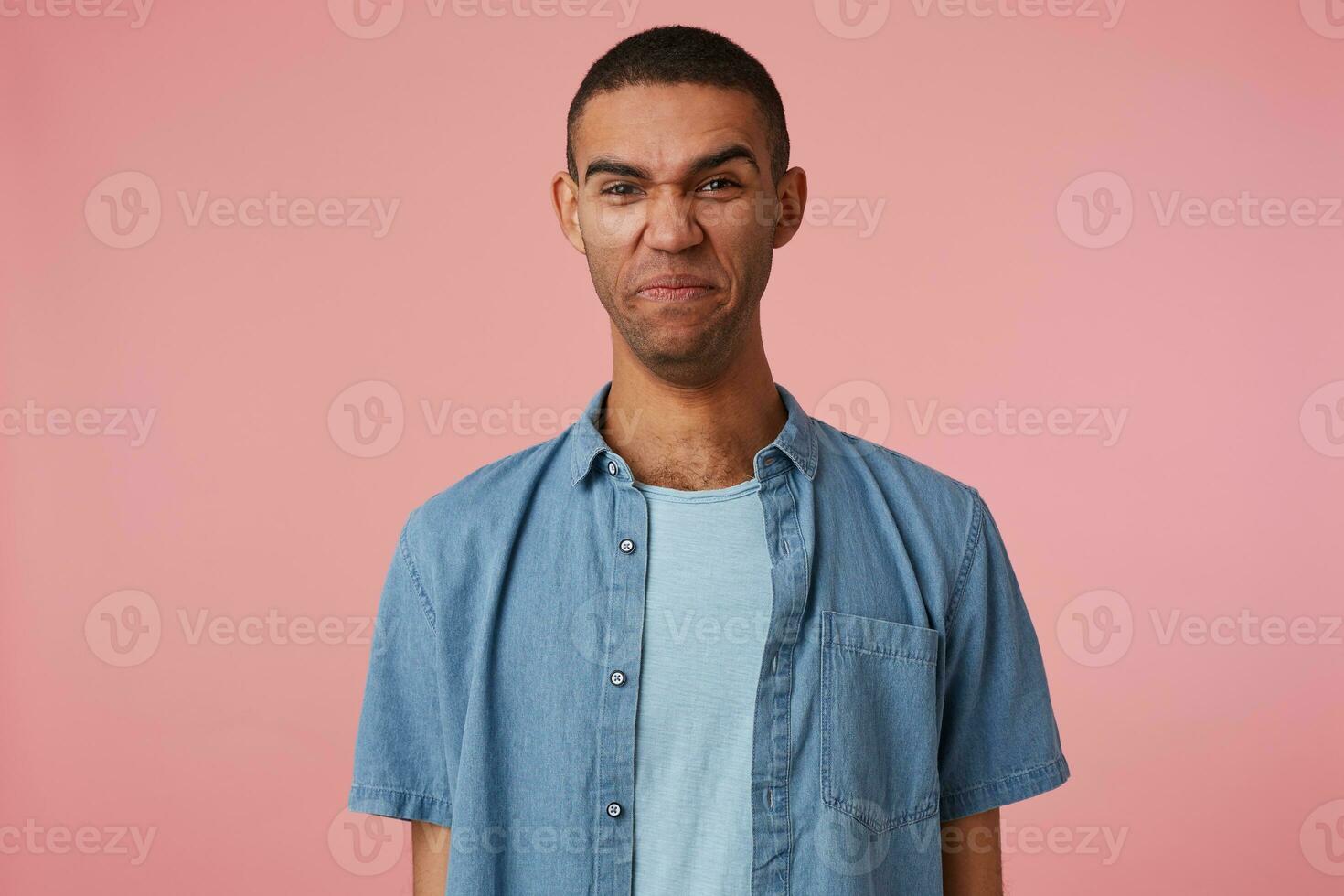 Photo of disgusted dark skinned boy with clenched lips, saw something repulsive and unpleasant and frowning, stands over pink background. People and emotion concept.