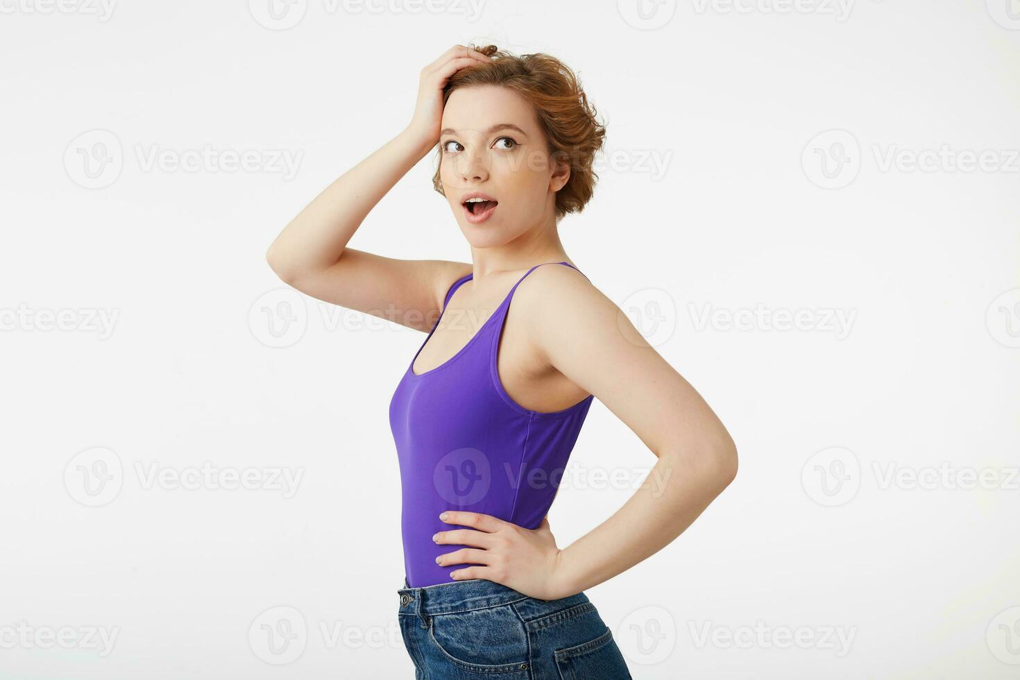 Portrait of hocked attractive short-haired girl, wearing a purple jersey, looking away in surprise with wide open mouth and eyes, straightens hair with his hand, isolated over white wall. photo