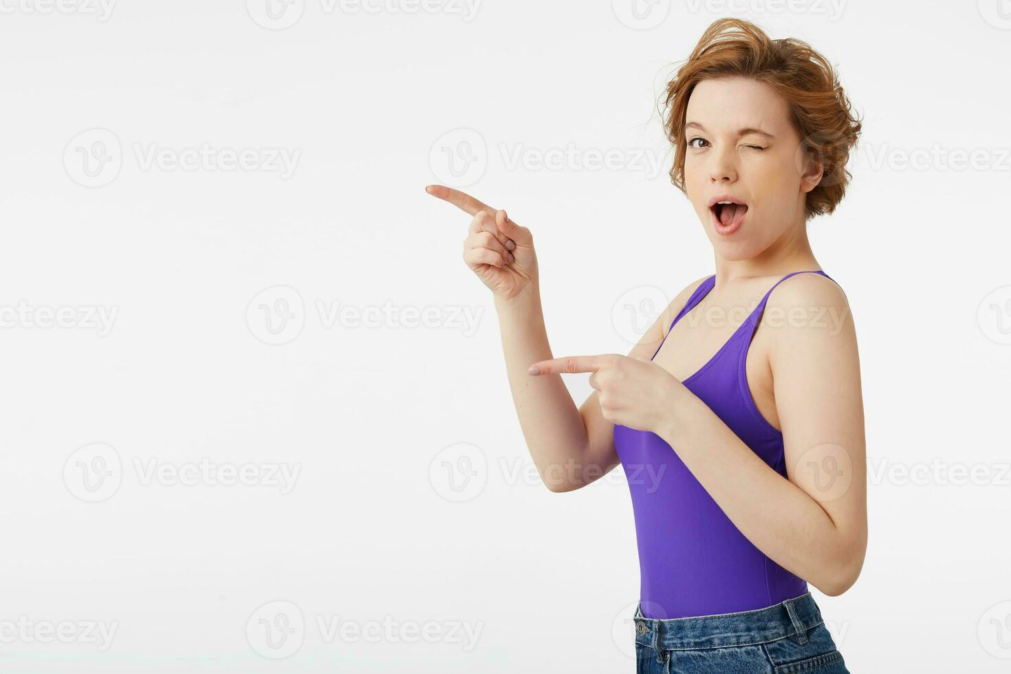 retrato de un joven atractivo de pelo corto chica, vistiendo un púrpura jersey y vaqueros, sonriente en general, parpadeo y mirando a el cámara, puntos el dedo a Copiar espacio aislado terminado blanco pared. foto