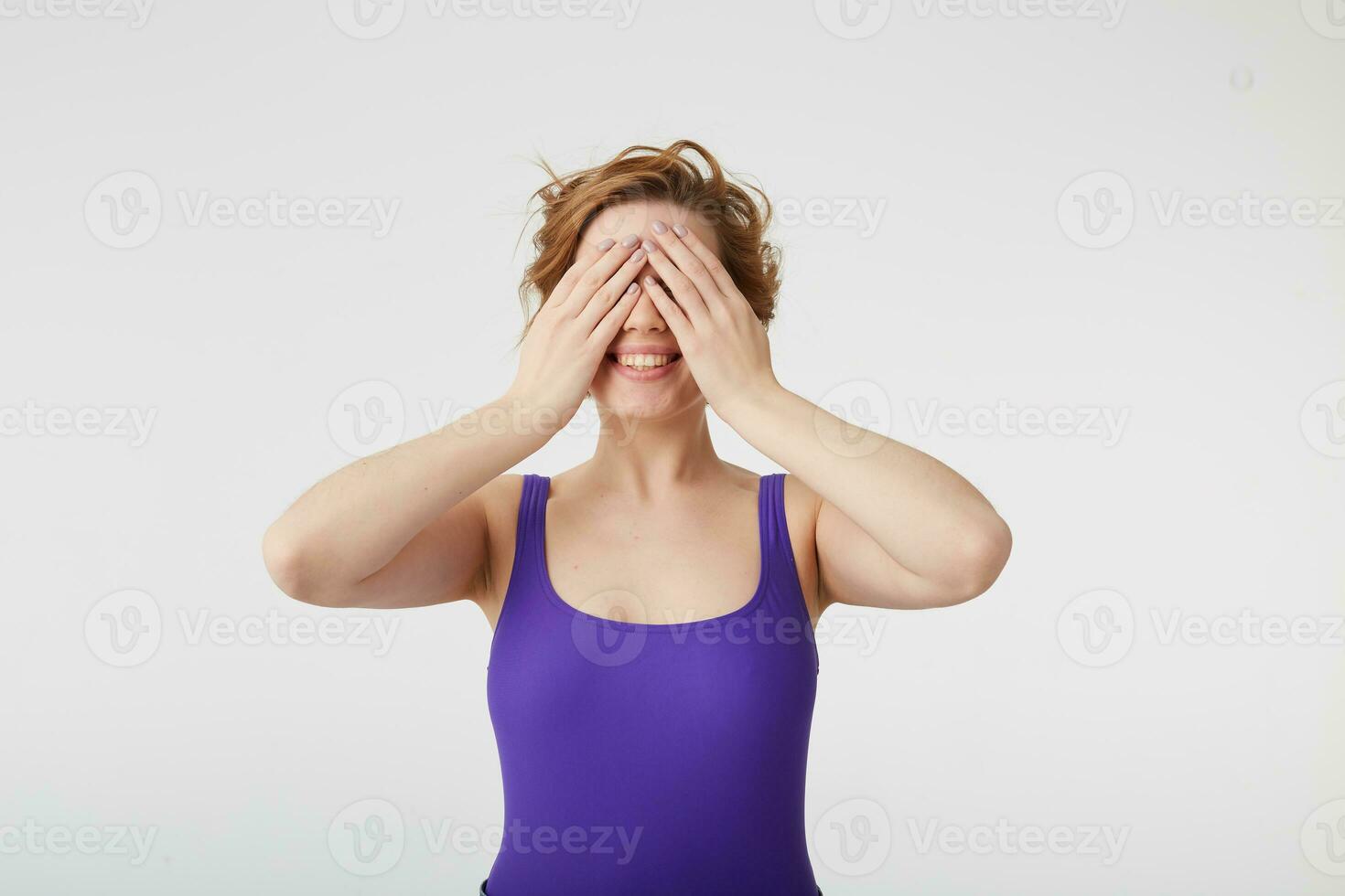 Cheerful lovely amazed young attractive short-haired girl with happy expression, covers eyes with hand, anticipates surprise, smiles broadly, dressed in casual clothes, stands over white wall. photo