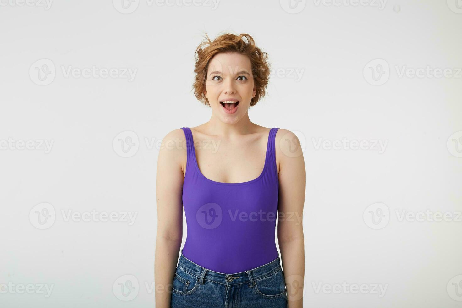 Photo of amazed young attractive curly short-haired girl, heard the incredible news and standsw with wide open mouth in surprise, stands over white wall.