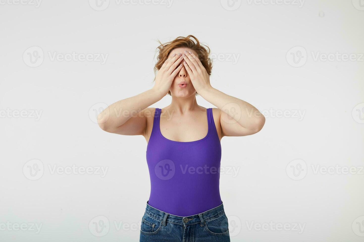 Happy amazed young attractive short-haired girl covers her eyes with hands going to see surprise prepared by her boyfriend. Young cheerful lady covering her face with hands stands over white wall. photo