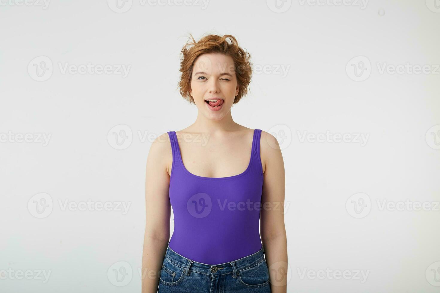 Photo of happy funny young cute short-haired girl, winks and shows tongue, wears in purple shirt, stands over white background.