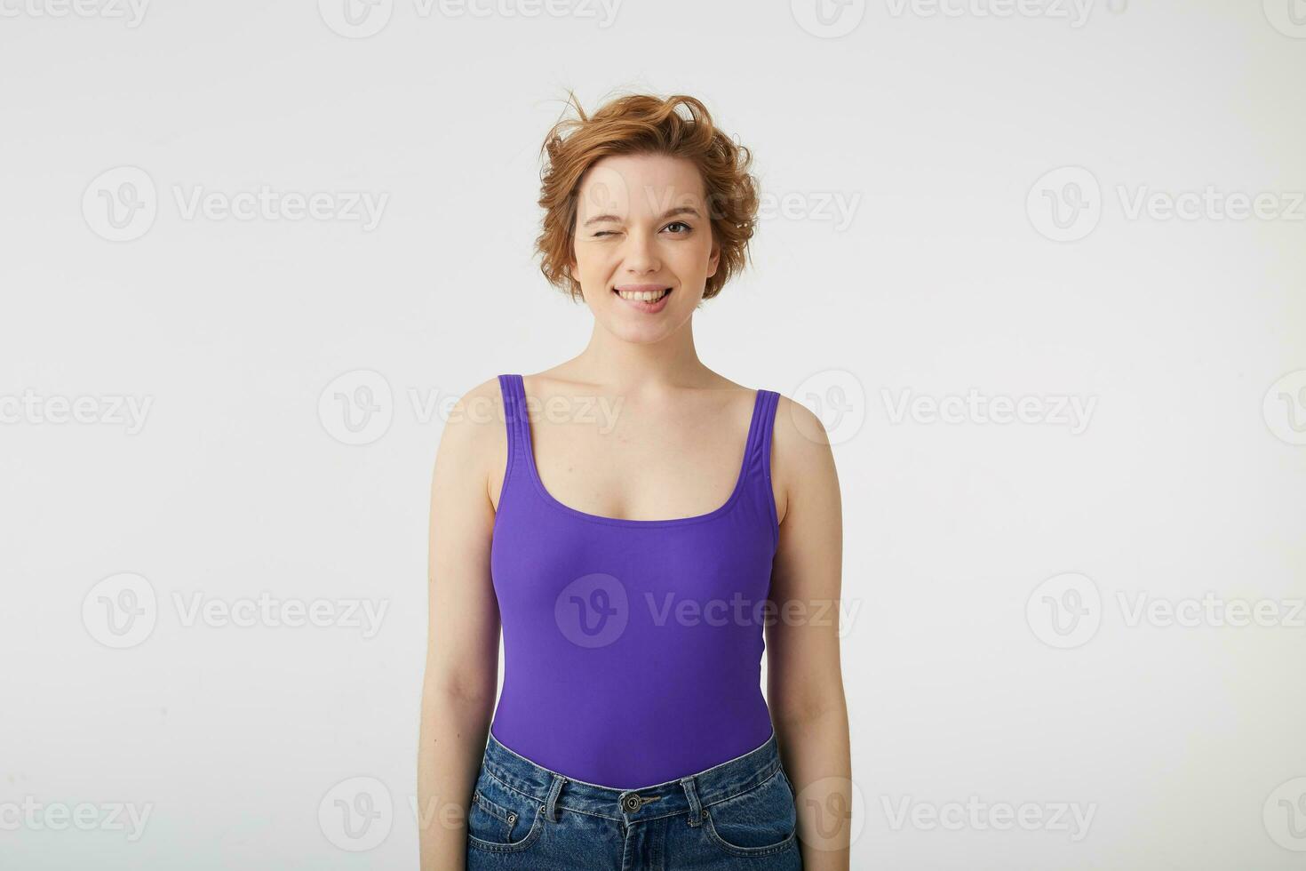 Portrait of happy funny young attractive short-haired girl, winks and bites lip, wears in purple shirt, stands over white background. photo