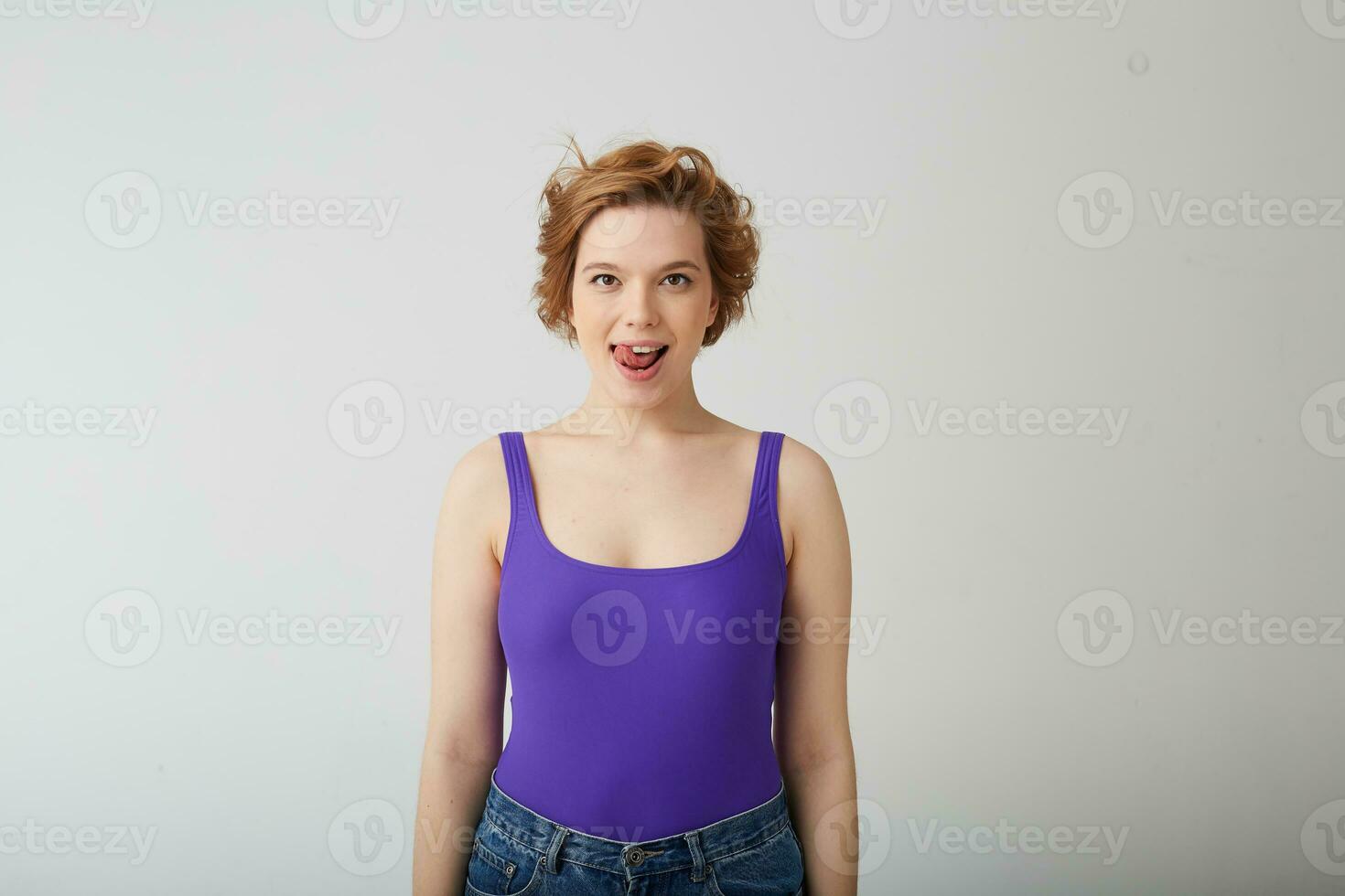 Photo of happy funny young cute short-haired girl, looking at the camera and licking, presenting a delicious dessert, stands over white background.