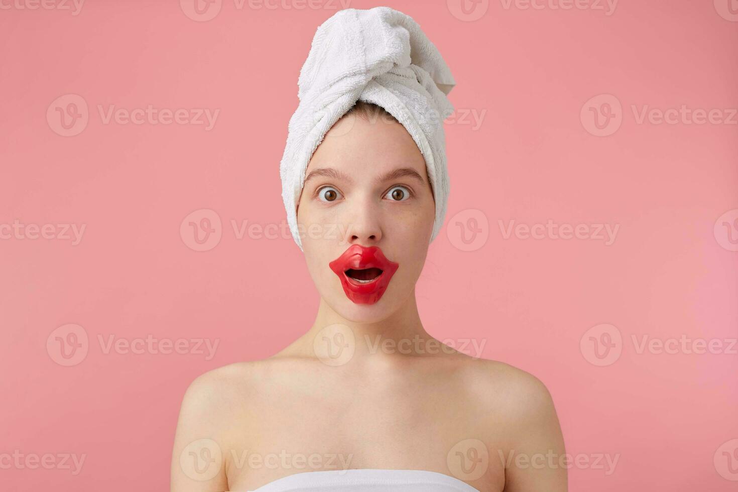 Close up of young dazed lady after spa with a towel on her head, looks at the camera, with wide open eyes and mouth, patch for lips, stands over pink background. photo