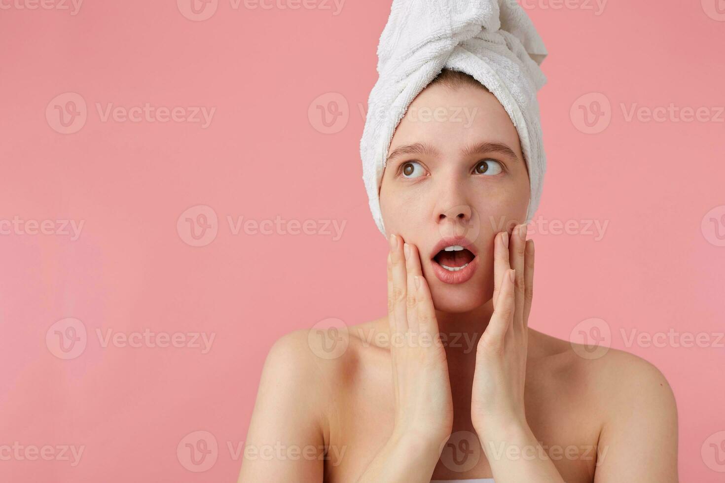 Photo of wondered young woman after shower with a towel on her head, looks away with wide open mouth and eyes, touches her cheeks, stands over pink background.