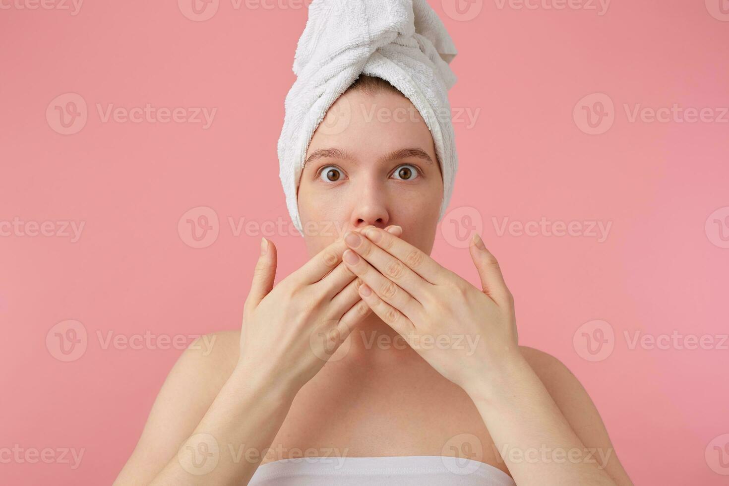 Photo of wondered young woman after shower with a towel on her head, covering mouth with hands, stands over pink background.