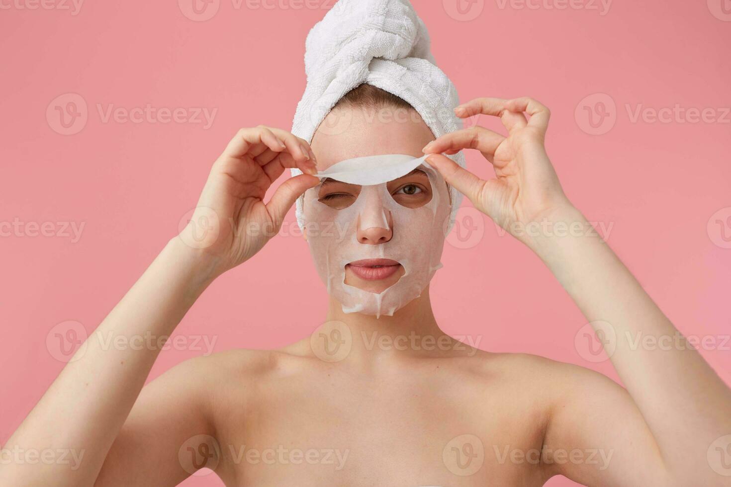 Close up of young woman with a towel on her head after shower, trying to remove the fabric mask from the face, stands over pink background. photo