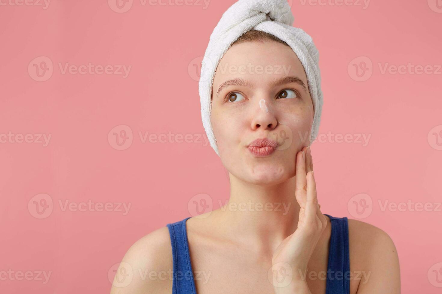 Photo of thinking young woman with a towel on her head after shower, can't make a decision, looks away and touches cheek, stands over pink background.