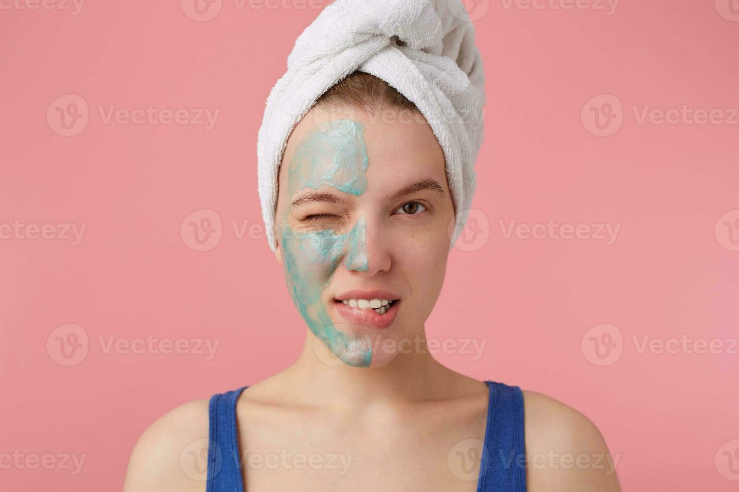 Portrait of young nice winked girl with half face mask, with a towel on her head after shower,smiling and looking at the camera over pink background. photo