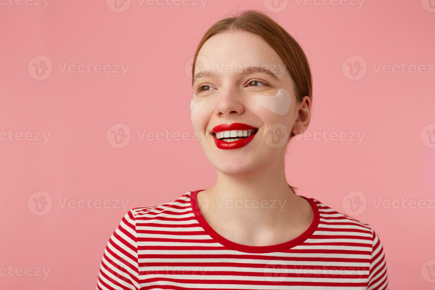 Close up of attractive young smiling red-haired woman with red lips and with patches under the eyes, wears in a red striped T-shirt, looks away with a happy expression, stands over pink background. photo