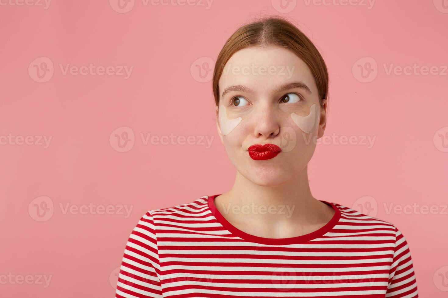 young mysterious red-haired lady with red lips and with patches under the eyes, wears in a red striped T-shirt, confused looks at the left side, something plotting, stands over pink background. photo