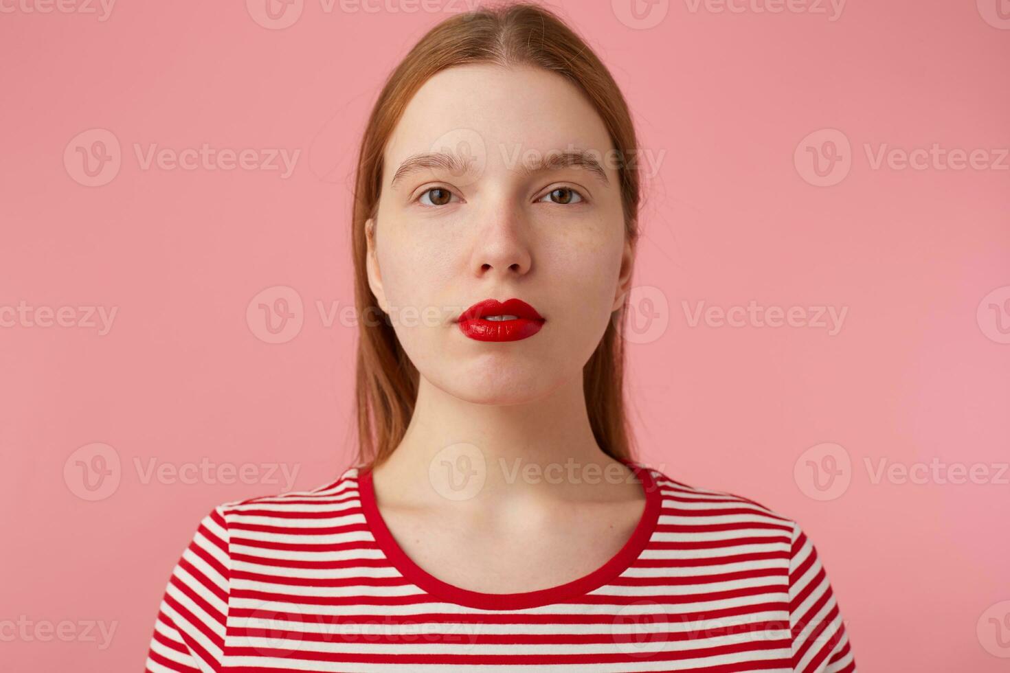 Close up of attractive young ginger woman with rad lips, wears in a red striped T-shirt, looks at the camera with a calm expression, stands over pink background. photo