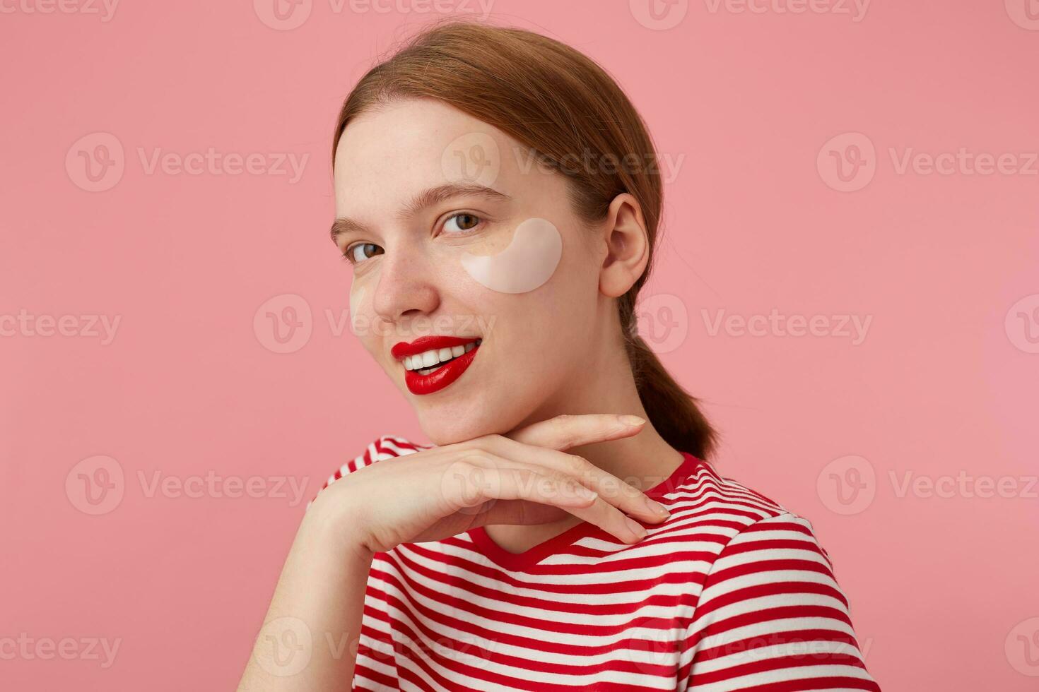 Nice smiling red-haired girl wears in a red striped T-shirt, with red lips and with patches under the eyes, touches the cheen, stands over pink background and enjoying free time for skin care. photo