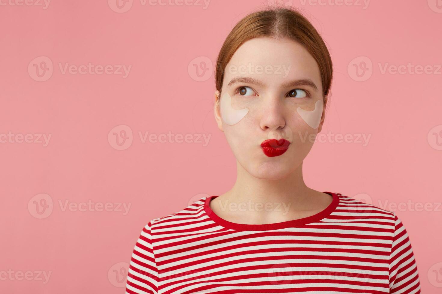 attractive young mysterious red-haired woman with red lips and with patches under the eyes, wears in a red striped T-shirt, looks at the left side, something plotting, stands over pink background. photo