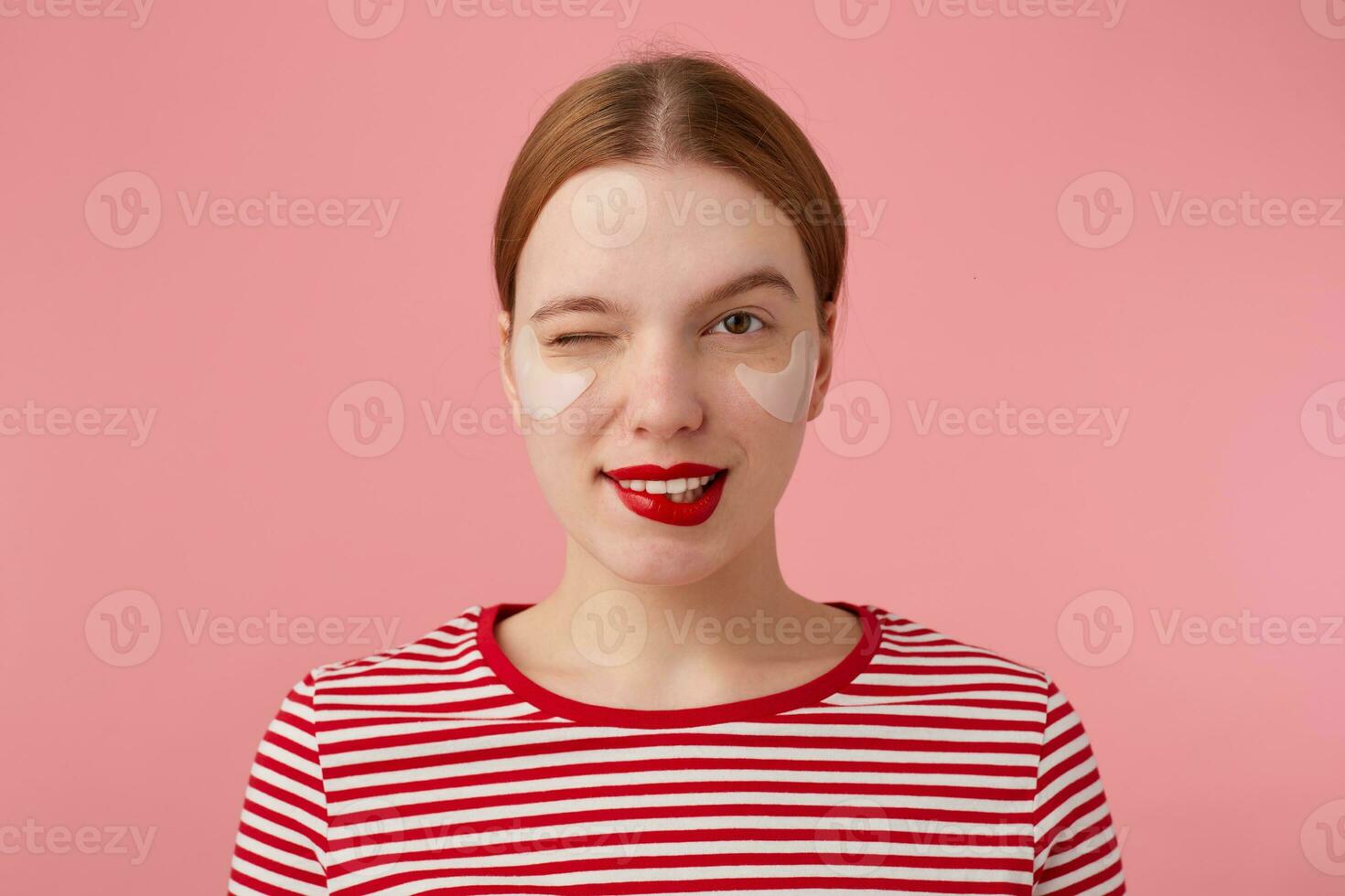 Portrait of cute young winking red-haired lady with red lips and with patches under the eyes, wears in a red striped T-shirt, looks at the camera and bites his lips, stands over pink background. photo