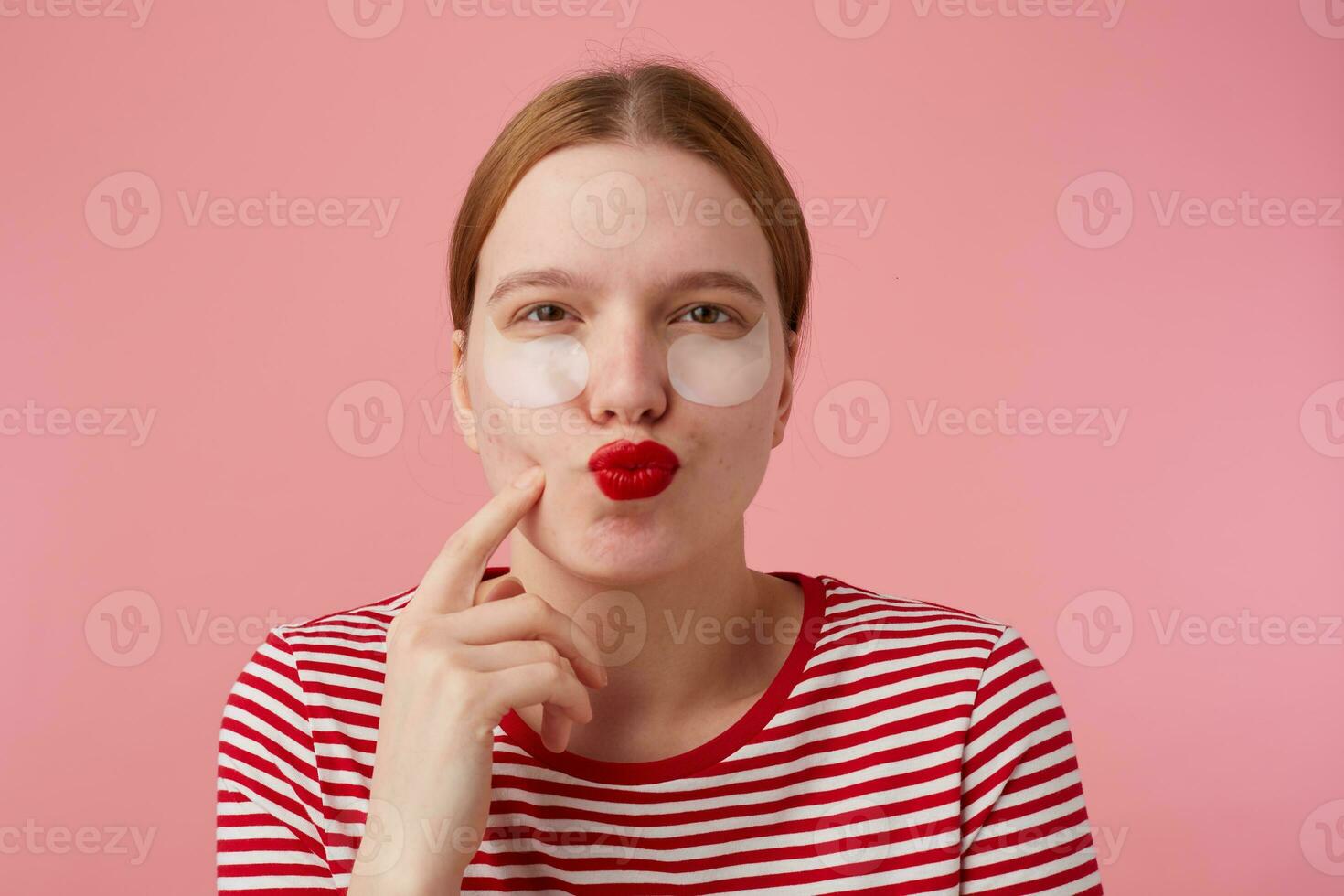 Portrait of thinking young red-haired girl with red lips and with patches under the eyes, wears in a red striped T-shirt, stands over pink background, points to his cheek and lokking at the camera. photo