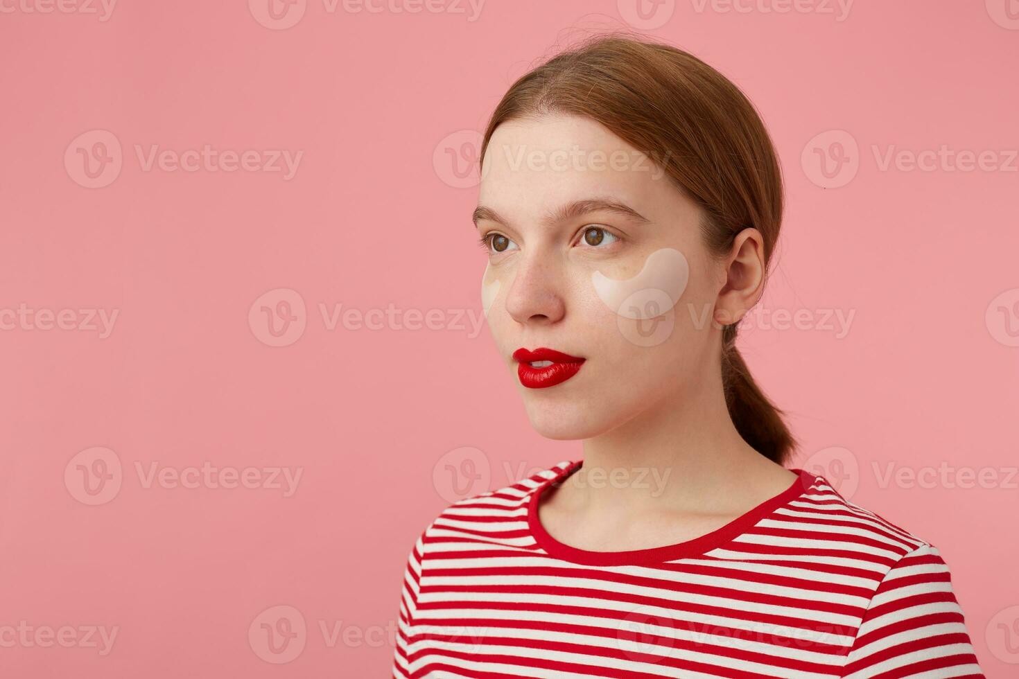 Close up of cute young ginger lady with rad lips, wears in a red striped T-shirt, looks at the camera with a calm expression, stands over pink background. photo