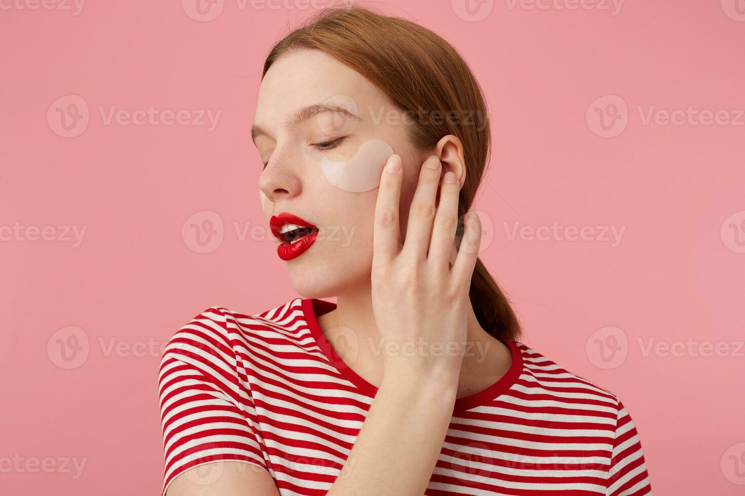 Young red-haired girl with red lips and with patches under the eyes, wears in a red striped T-shirt, touches the cheek, stands over pink background with closed eyes enjoying free time for skin care. photo