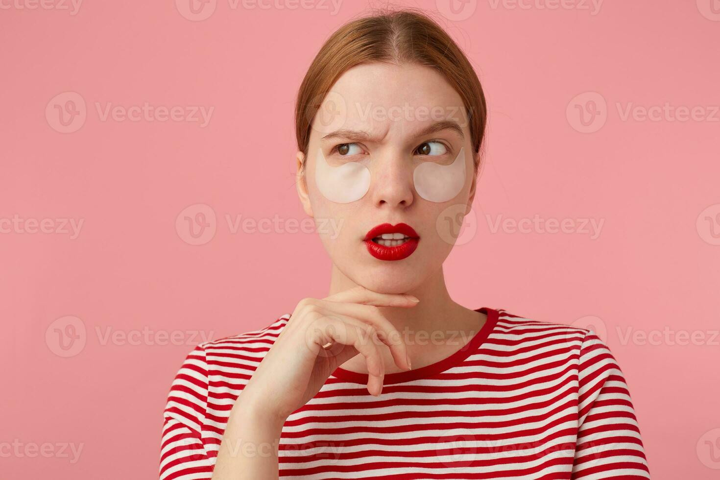 Cute thinking young red-haired girl with red lips and with patches under the eyes, wears in a red striped T-shirt, touches the cheek, looks away, stands over pink background. photo