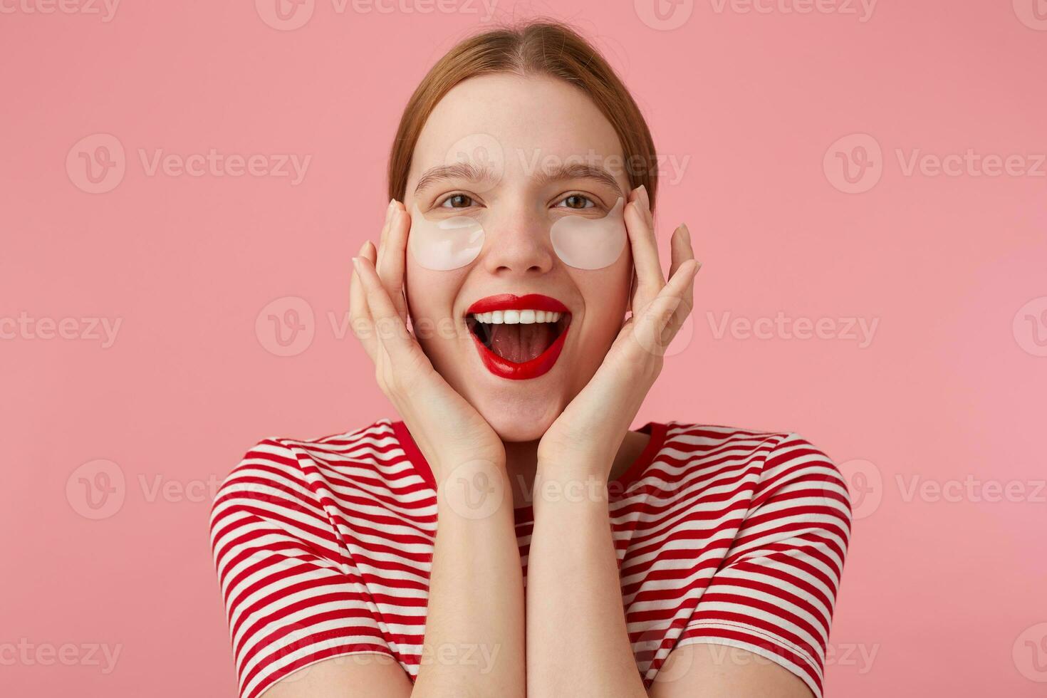 Nice young happy red-haired lady in a red striped T-shirt, with red lips , touches his face with fingers, very pleased with new patches. Stands over pink background. photo