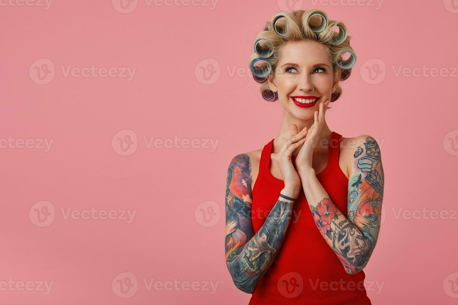 alegre joven atractivo rubia dama con tatuado manos sonriente y mirando soñando hacia arriba, anticipando próximo fiesta y preparando para él, en pie en contra rosado antecedentes foto