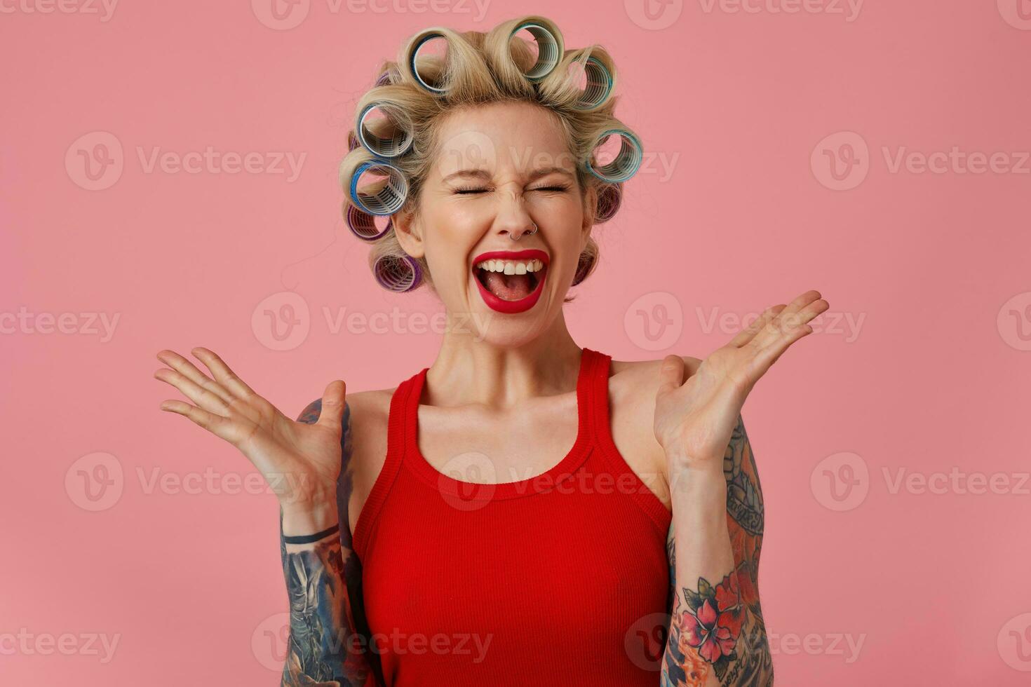 Overjoyed young blonde female with tattooes screaming happily with closed eyes and raising emotionally hands, having curlers on her head while posing over pink background photo