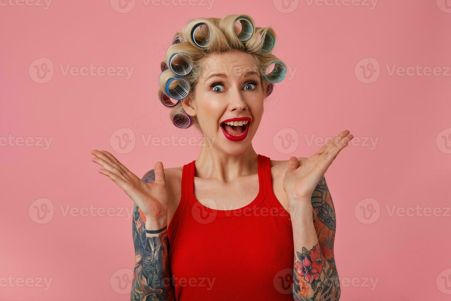 retrato de con los ojos abiertos atractivo joven tatuado dama curling su rubia pelo y vistiendo festivo maquillaje mientras posando terminado rosado fondo, mirando alegremente a cámara con amplio boca abrió foto