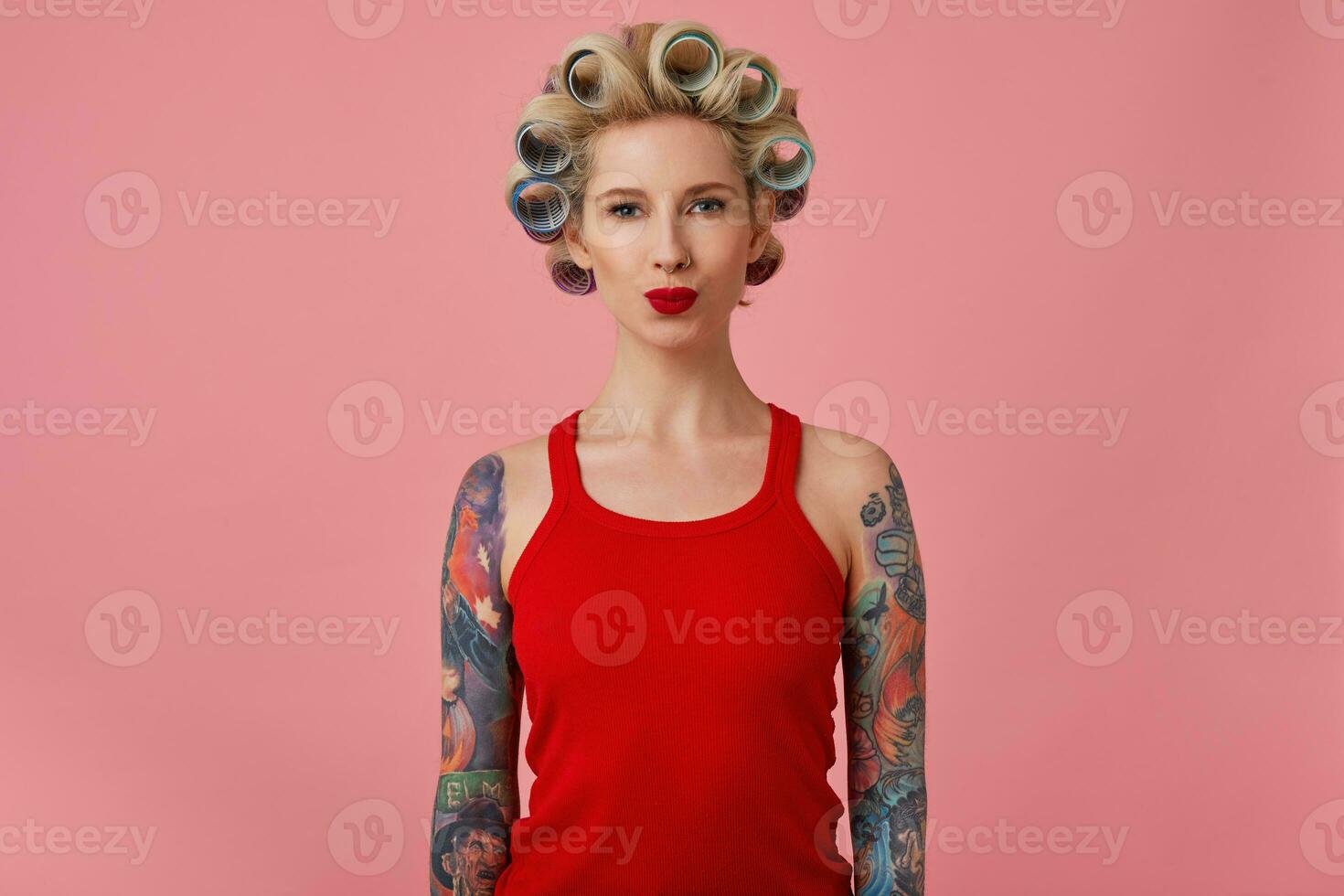 Portrait young attractive blonde woman with tattooes and festive makeup pouting her lips while looking positively at camera, having curlers on her head while standing over pink background photo