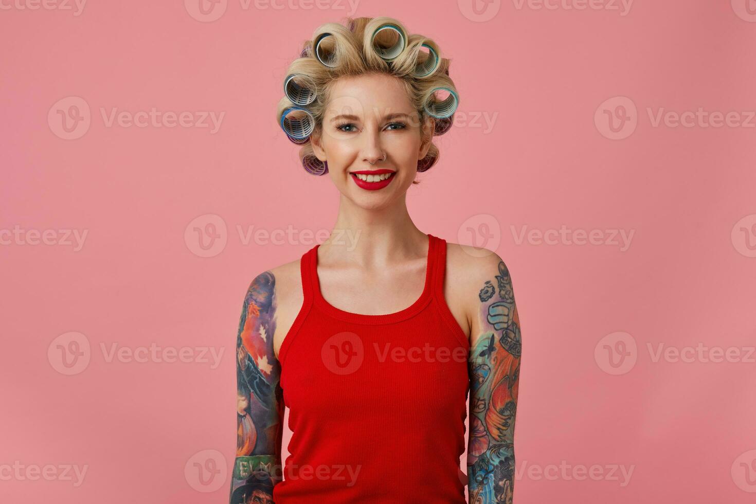 interior foto de joven positivo bonito rubia mujer con tatuajes teniendo rulos en su cabeza mientras en pie terminado rosado fondo, vistiendo festivo maquillaje y preparando para noche yendo fuera