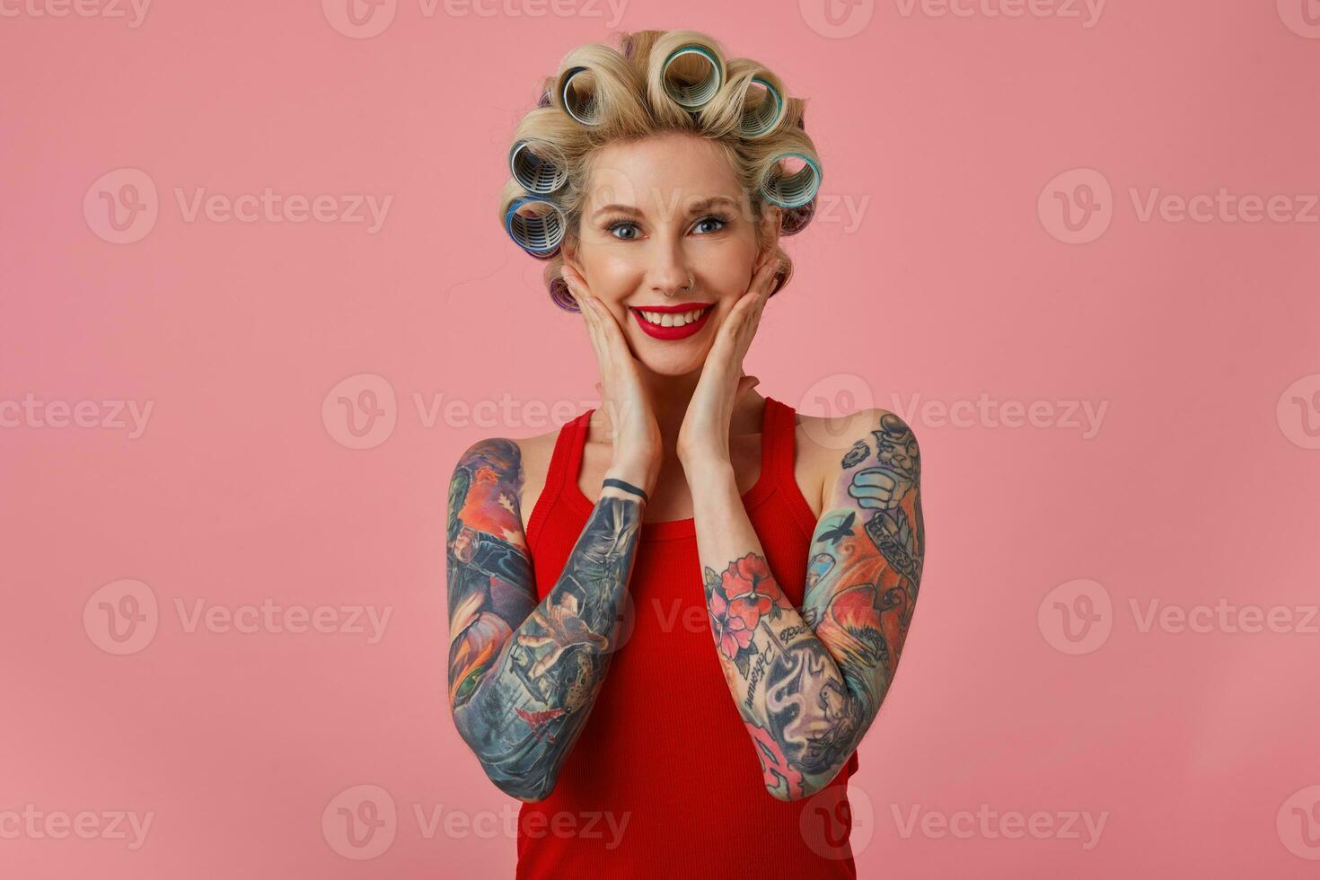 Portrait of beautiful young happy blonde female holding palms on her cheeks and smiling widely while posing over pink background, having tattooes on her hands and curlers on her head photo