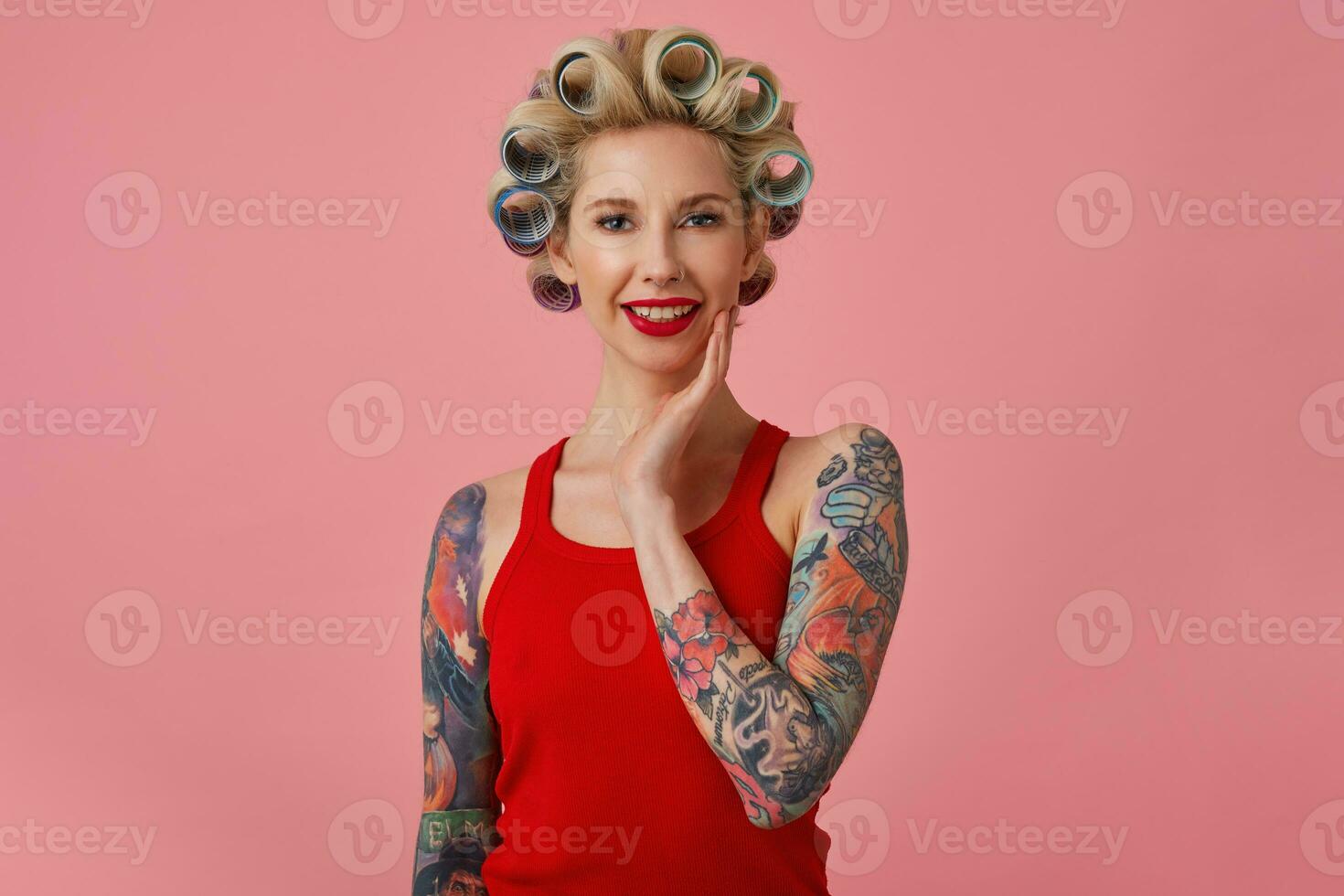 Charming young blonde tattooed woman with evening makeup making hairdo before upcoming party, touching soft her face and smiling gently at camera, isolated over pink background photo