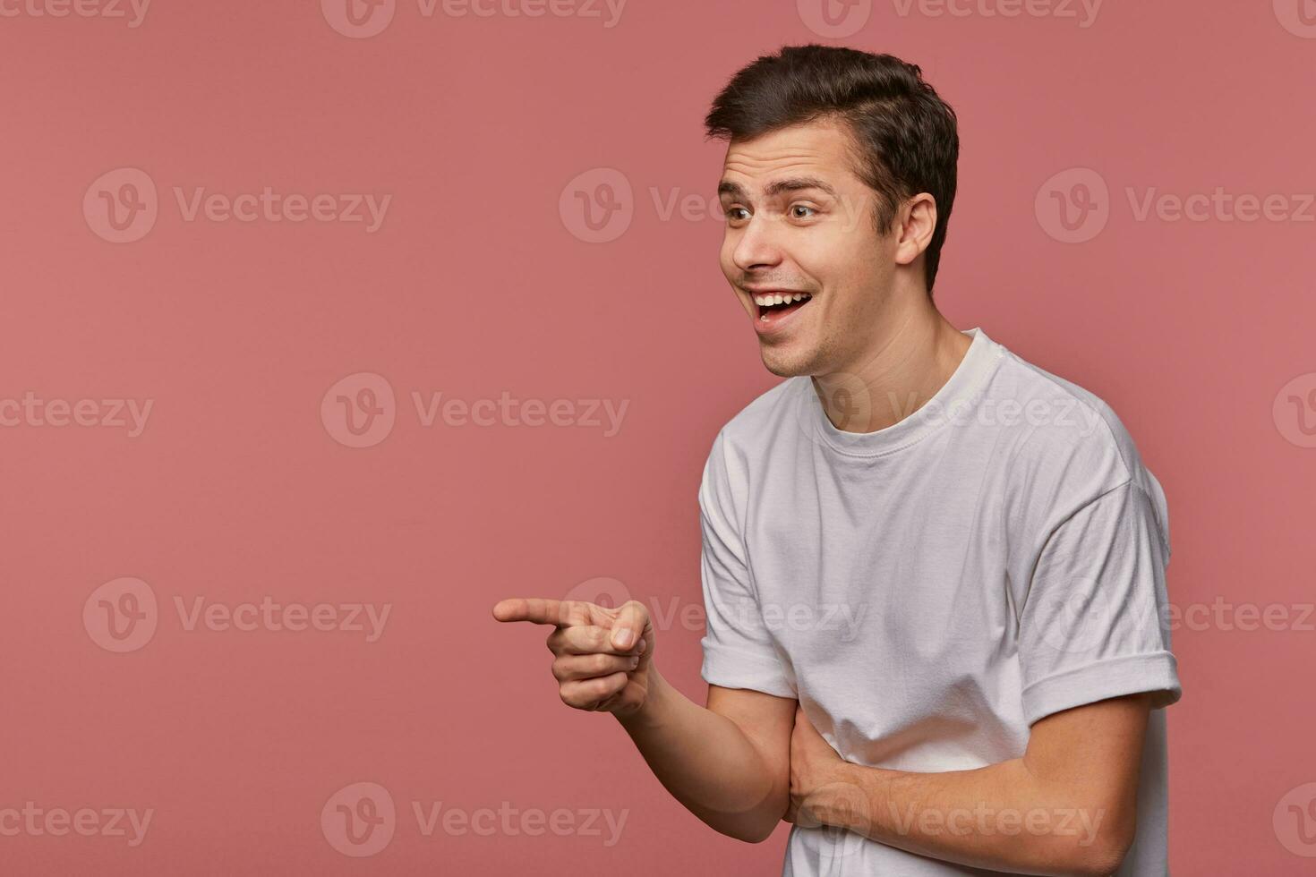 Indoor portrait of happy young dark haired male looking joyfully aside, raising hand and pointing with index finger, standing over pink background in grey t-shirt photo
