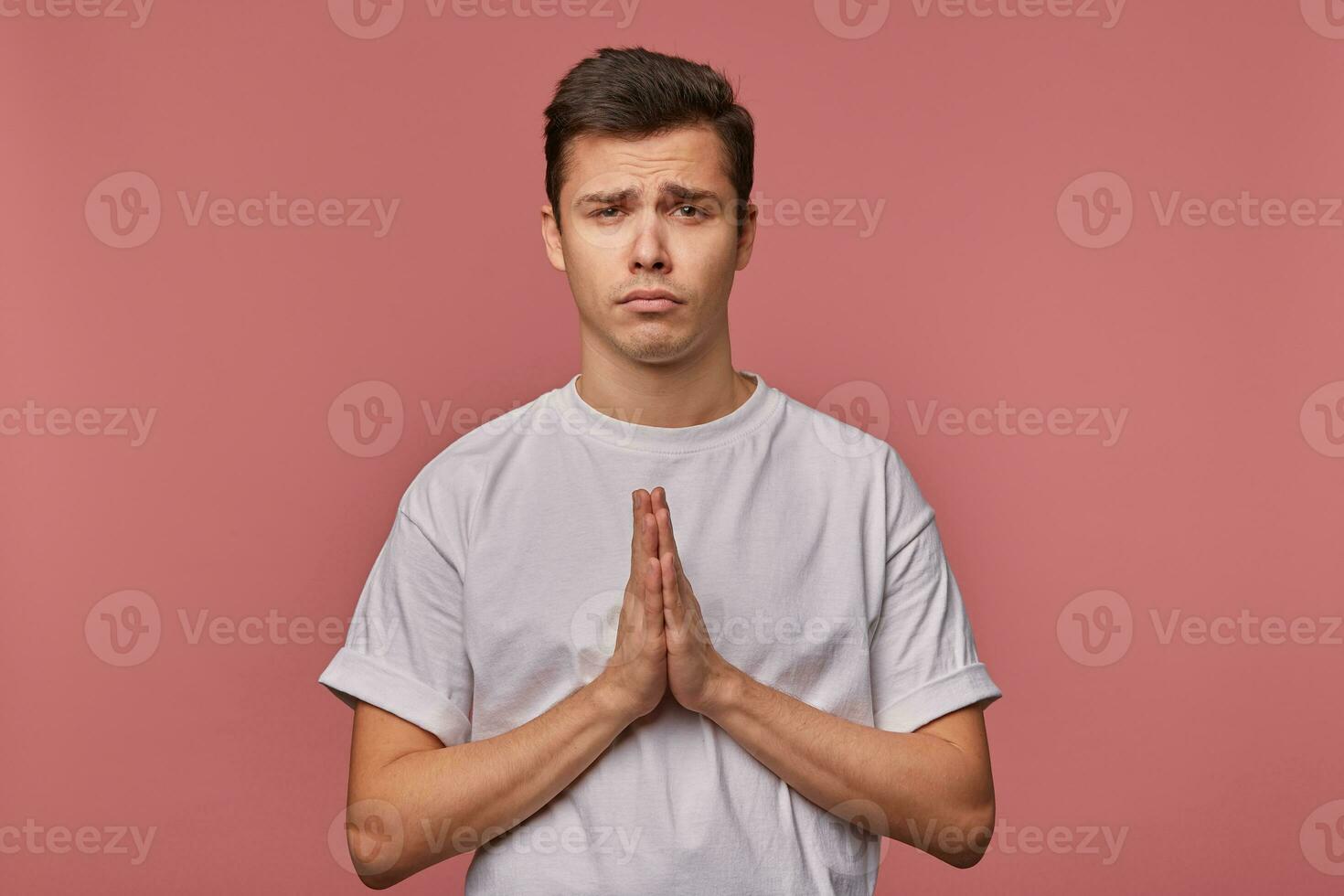 Studio shot of pretty dark haired young male with short haircut posing over pink background with raised hands, keeping palms together and begging for something pitifully photo