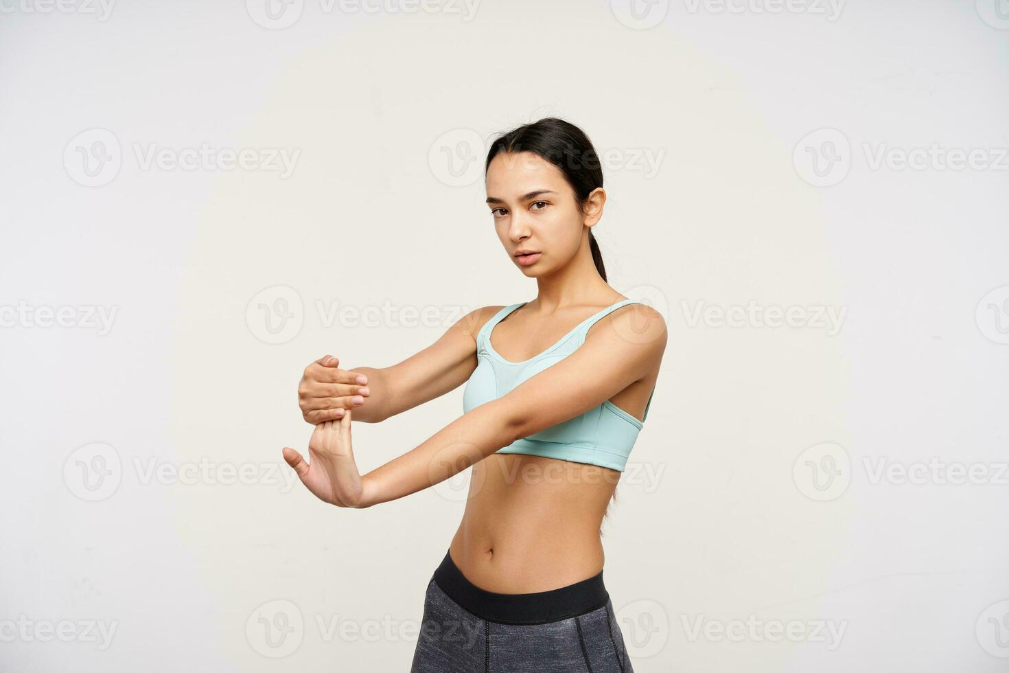 Indoor shot of attractive young brown haired woman pulling her fingers while stretching muscules and looking attentively at camera, isolated over white background photo
