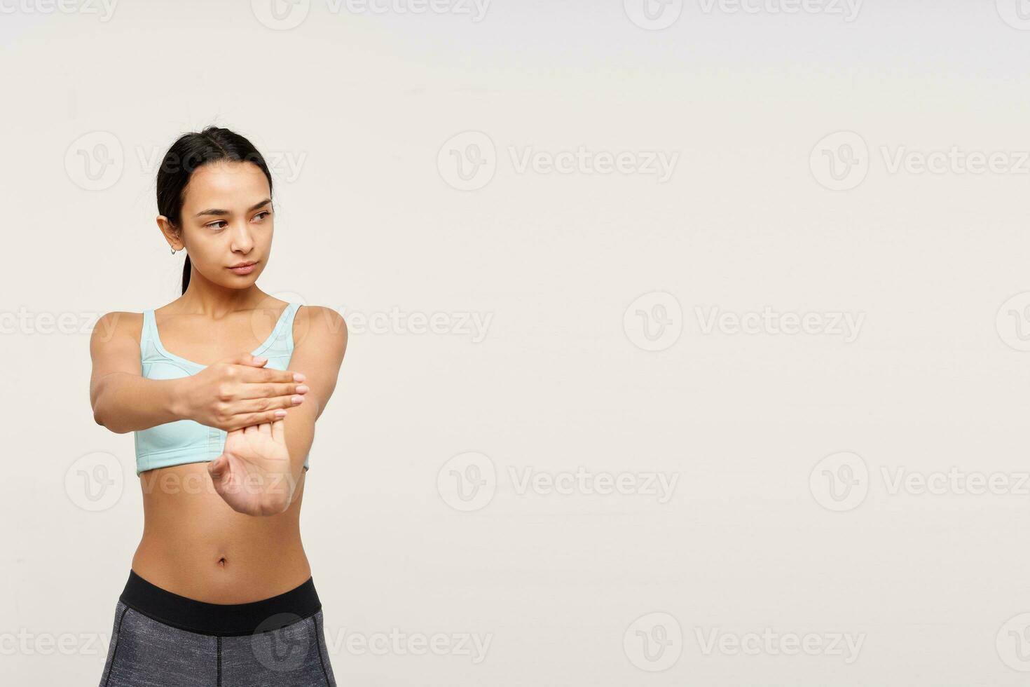 Studio shot of young brown-eyed woman without makeup keeping her hands raised while stretching muscules, standing over white background in sporty wear photo