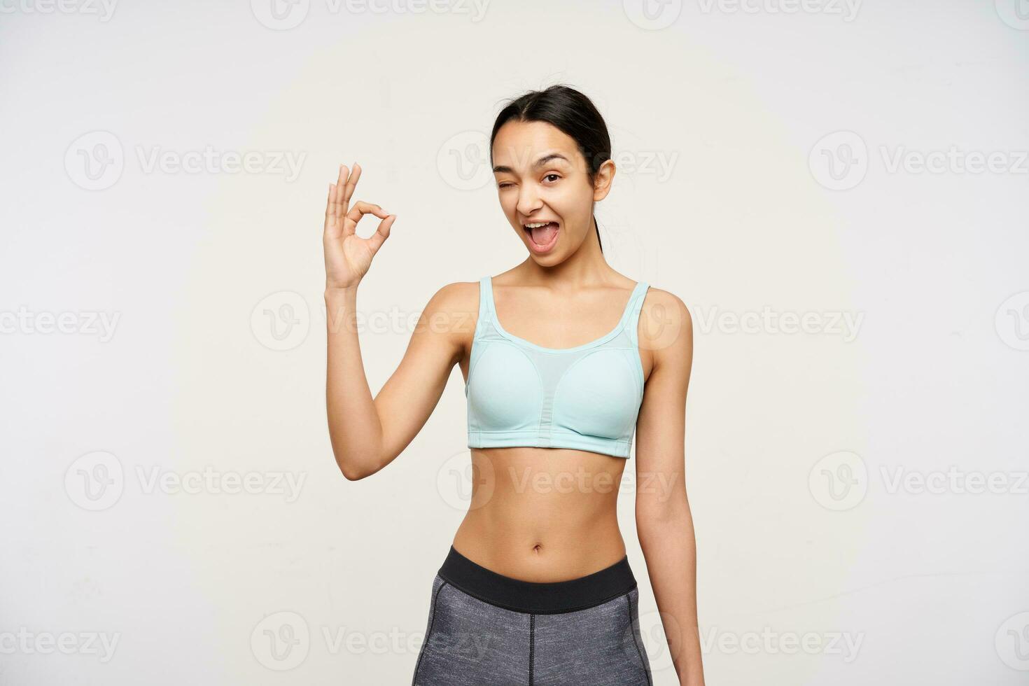 Joyful young lovely brown haired slim woman giving happily wink at camera while showing well done sign with raised hand, standing over white background photo