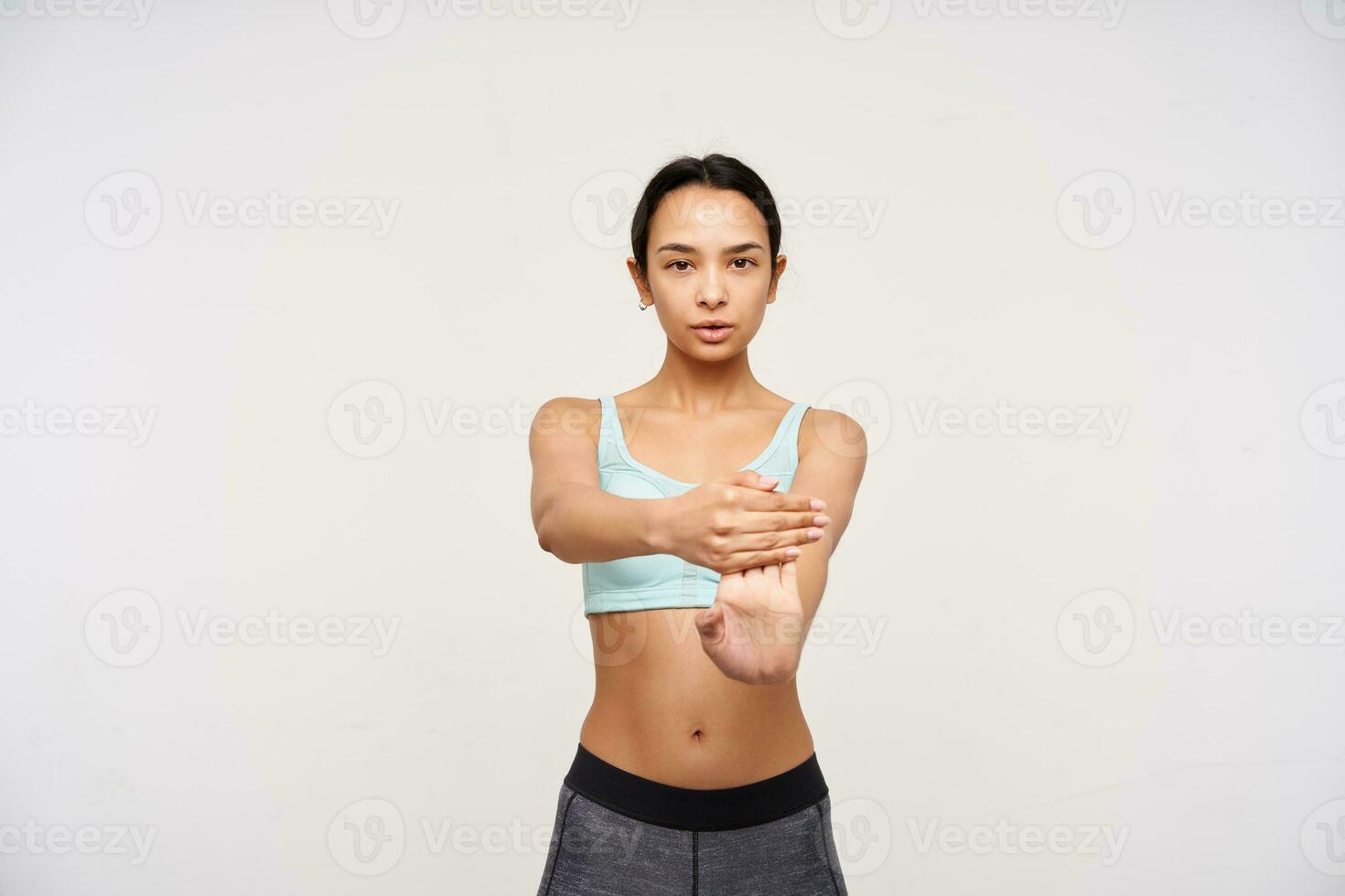 Serious young pretty brown haired lady with casual hairstyle being concentrated while training her hands, dressed in sporty bra and leggins while posing over white background photo