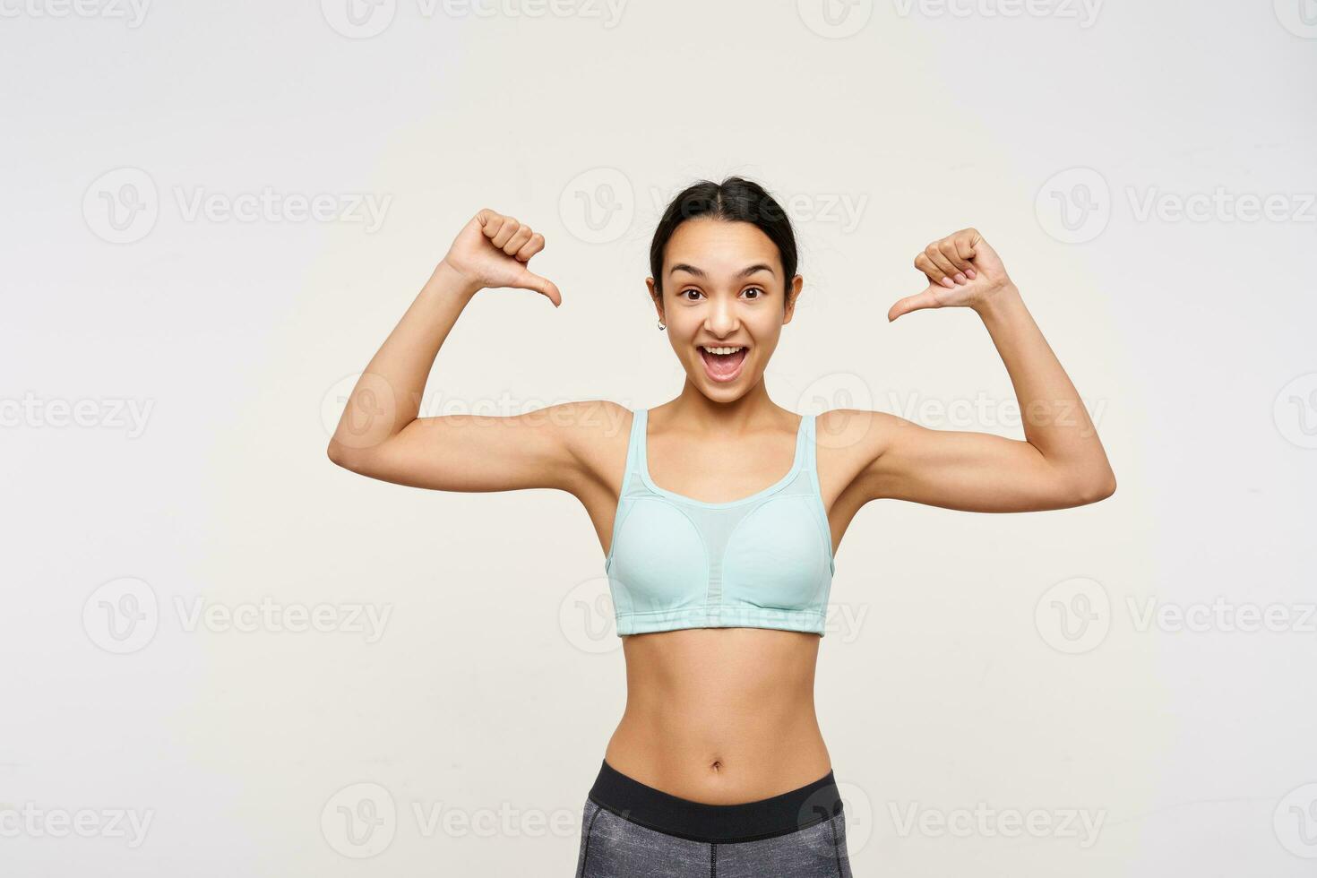 Excited young pretty brown haired sporty lady with casual hairstyle looking amazedly at camera while thumbing on herself, standing over white background photo