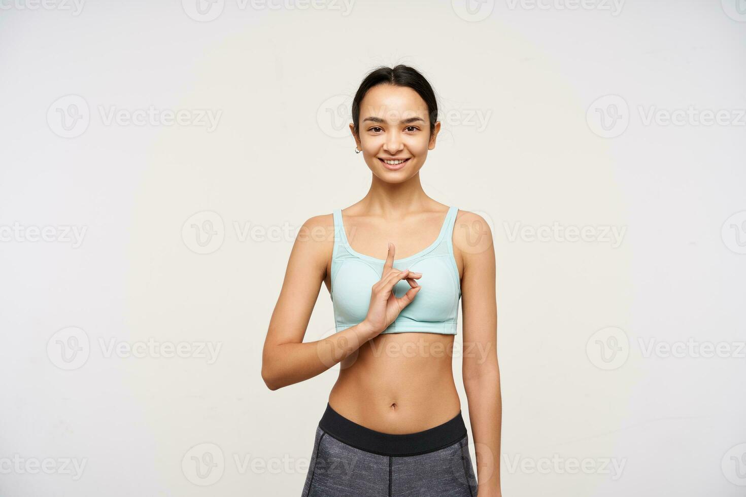 Cheerful young lovely brown haired lady raising hand with mudra gesture and looking positively at camera with broad smile, isolated over white background photo
