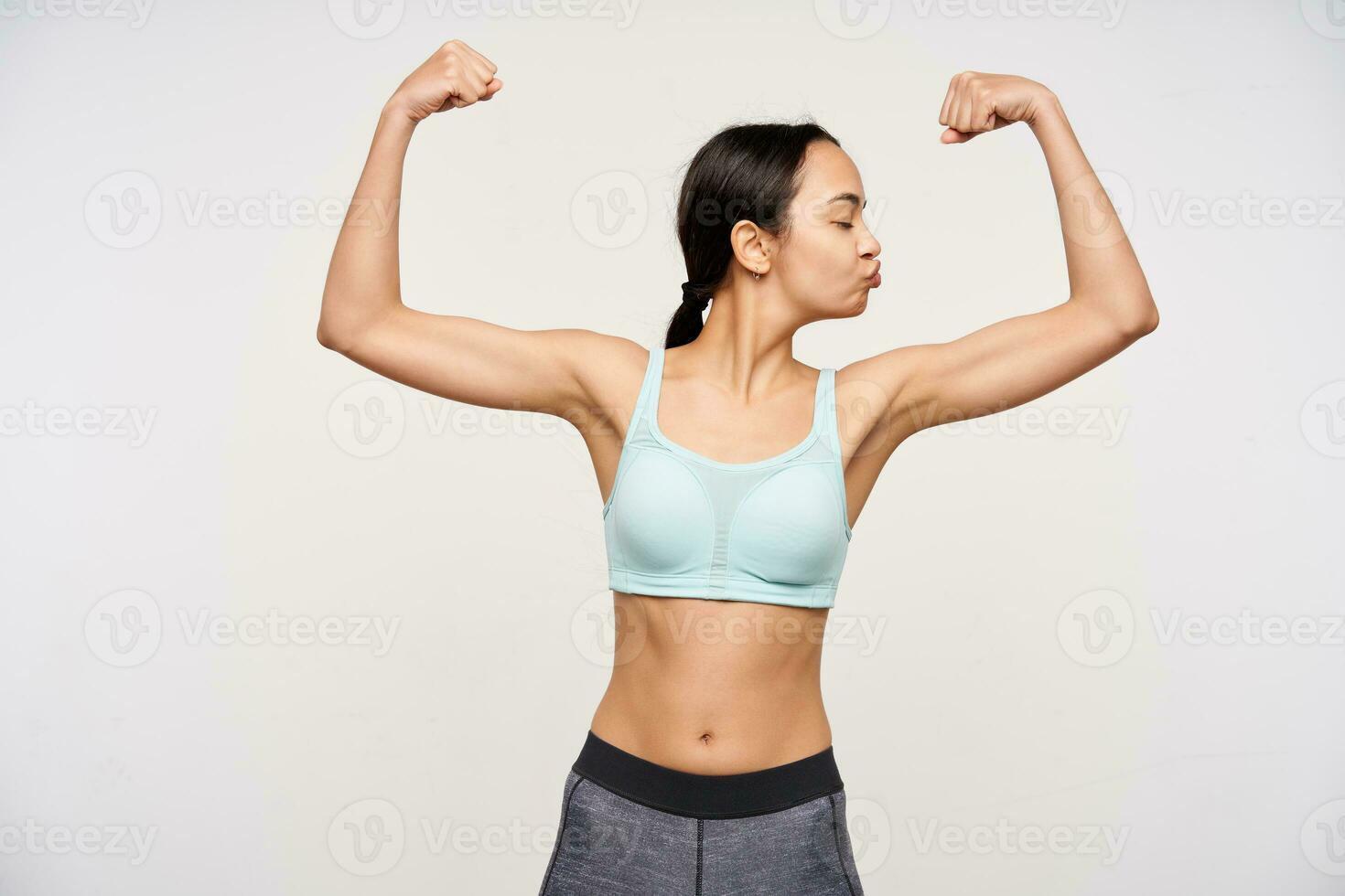 Good looking self-confident young brown haired lady keeping her eyes closed and folding lips in kiss while demonstrating her strong biceps, isolated over white background photo